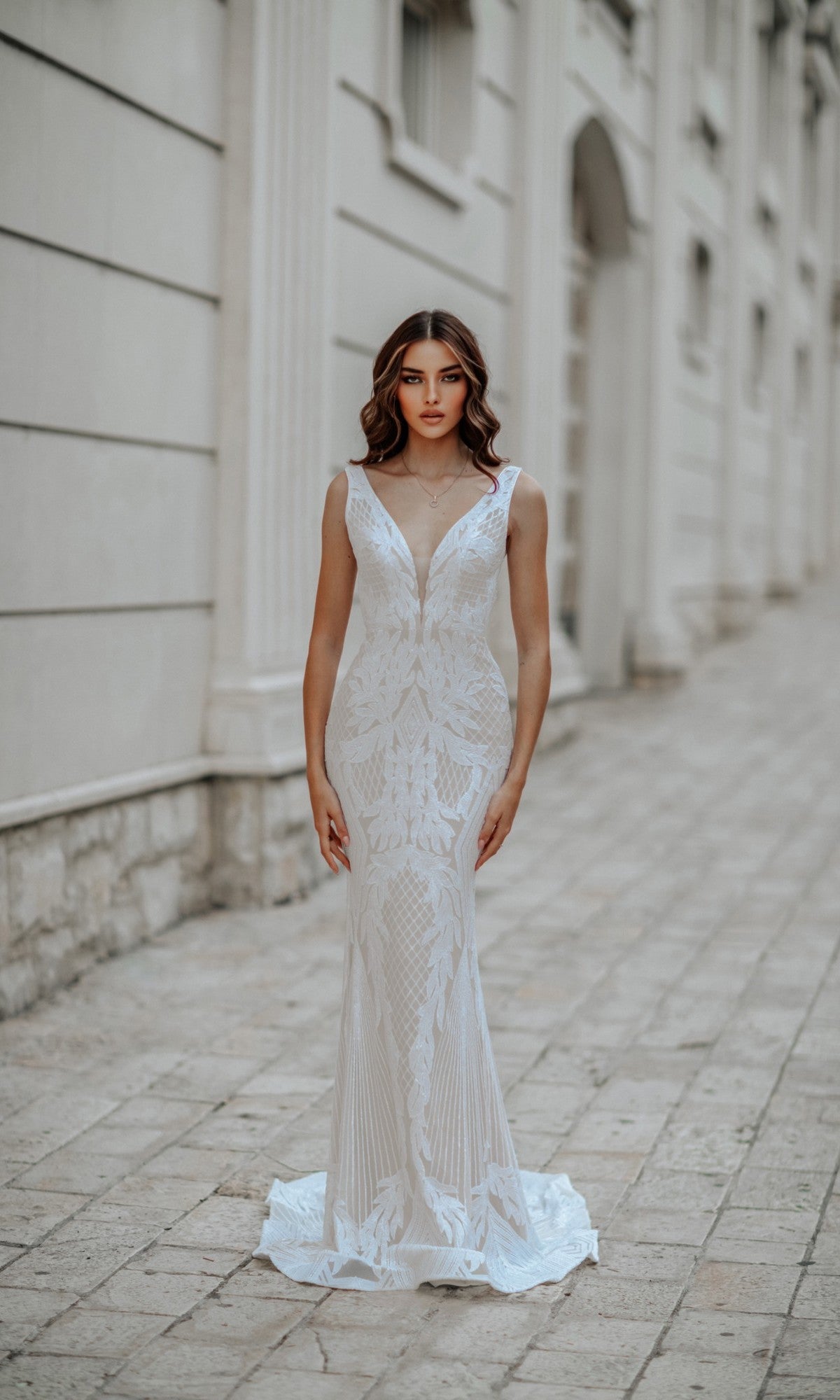 A woman wearing the Classic Long White Formal Dress: Tina Holly BB052, featuring a deep V-neckline and fitted silhouette, stands on a stone-paved walkway in front of a light-colored building.